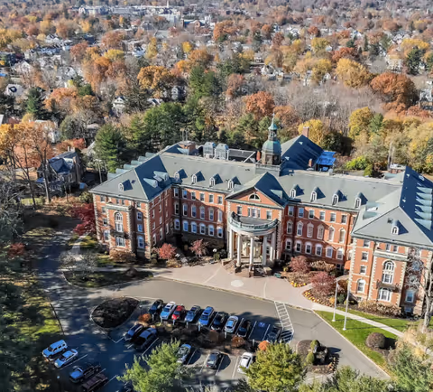Aerial view of a large, historic brick building with a central entrance featuring columns and a cupola on the roof. The building is surrounded by trees with autumn foliage and a parking lot with several cars parked. Residential houses and more trees are visible in the background.