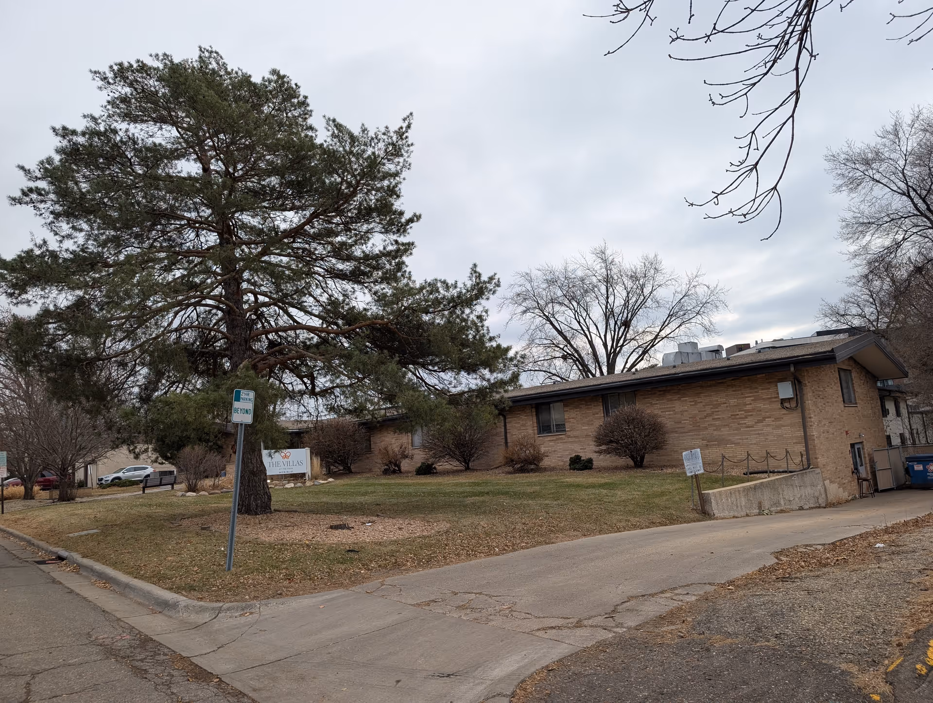 Exterior view of a single-story brick building with a sloped roof, surrounded by leafless trees and a large pine tree on a cloudy day. There is a sign on the lawn that reads 'The Villas at the Park' and a parking sign near the sidewalk.