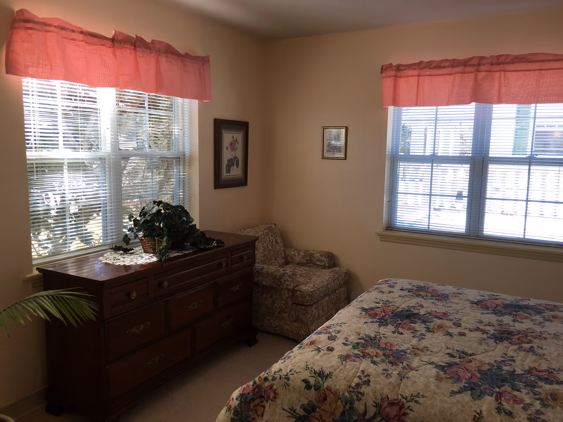 A bedroom with two windows covered by pink valances and white blinds. There is a wooden dresser with a potted plant on top, a floral upholstered armchair in the corner, and a bed with a floral patterned bedspread.