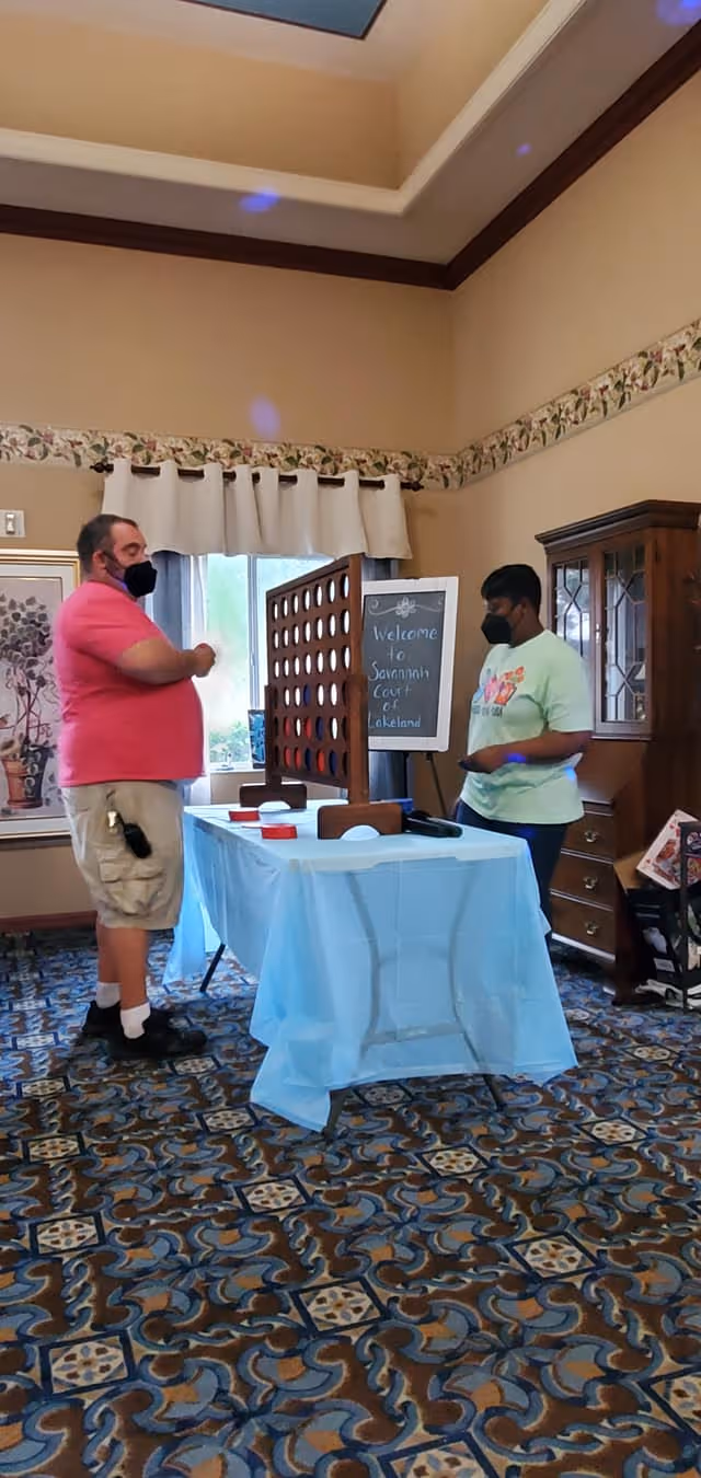 Two people wearing face masks playing a large Connect Four game on a table covered with a light blue tablecloth in a room with patterned carpet and beige walls. A sign in the background reads 'Welcome to Savannah Court of Lakeland.'