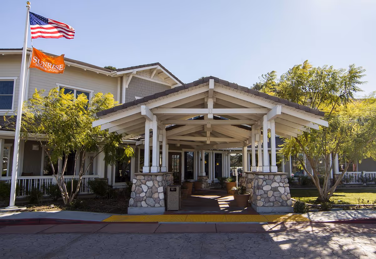 Covered entrance and porte-cochere of a senior living building with stone pillars, landscaping and American and Sunrise flags.