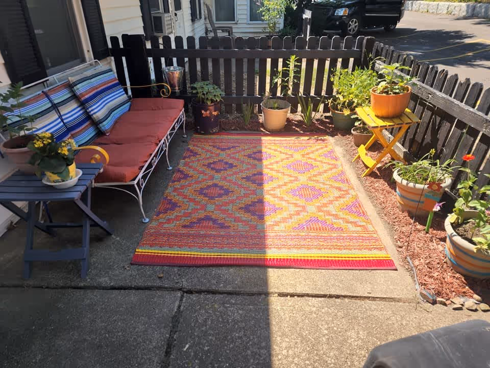 A small outdoor patio area with a colorful patterned rug on the concrete floor, a white metal bench with red cushions and striped pillows, a small blue side table with a potted yellow flower, and several potted plants arranged along a wooden fence. A yellow folding table holds a large orange pot with a plant. A black vehicle and part of a driveway are visible beyond the fence.