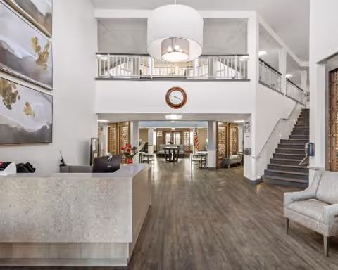 Bright two-story senior living facility lobby with a reception desk, seating, staircase and an upper balcony overlooking the entry.