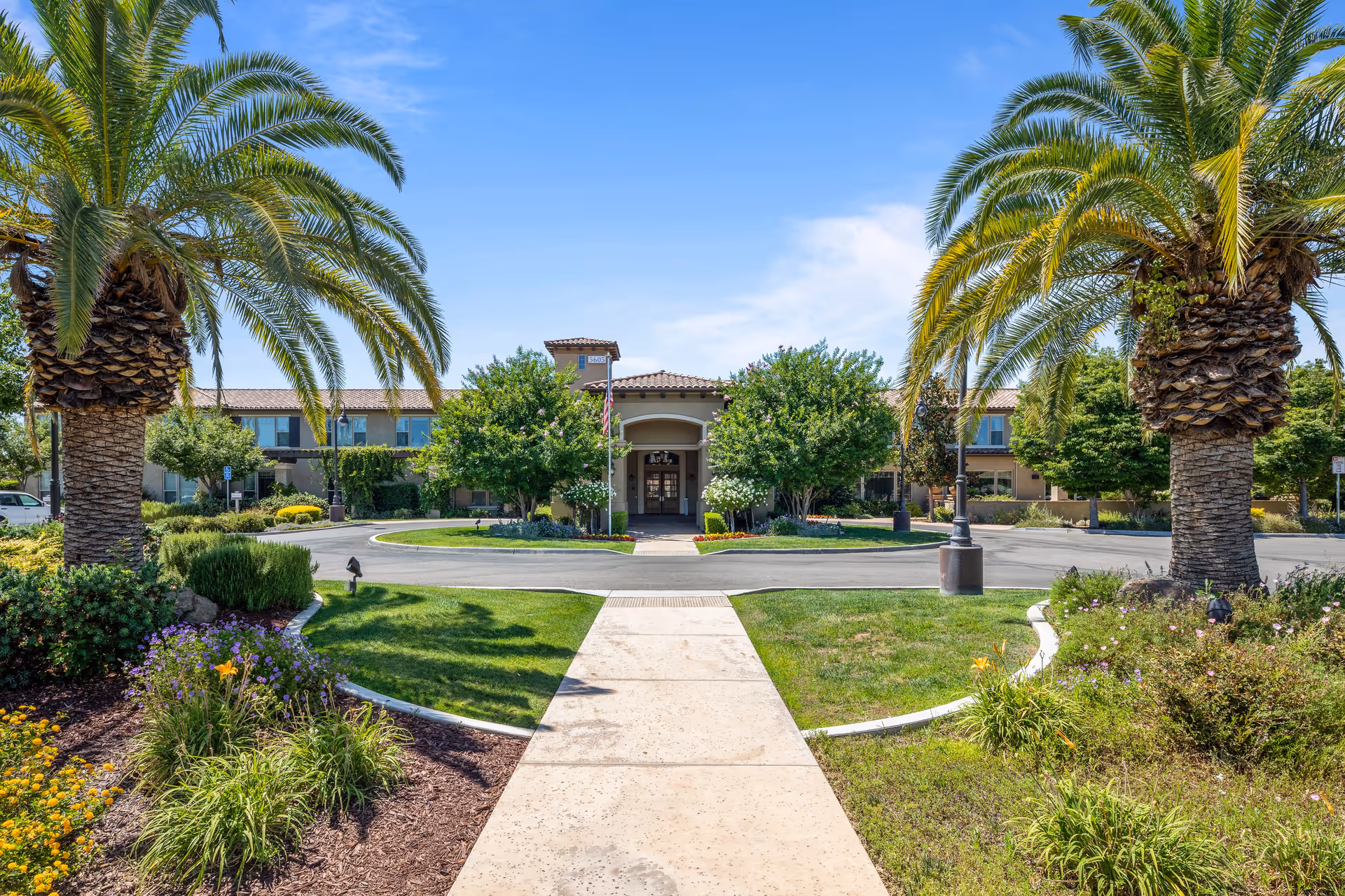 View of the front exterior of Oakmont of Fresno senior living facility with a paved walkway leading to the entrance, surrounded by palm trees, green lawns, and landscaped flower beds under a clear blue sky.
