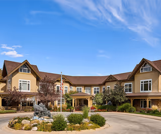 Exterior view of a two-story assisted living facility with beige siding and brown roof. The building is surrounded by landscaped greenery, trees, and a circular driveway with a statue in the center. The sky is clear and blue.