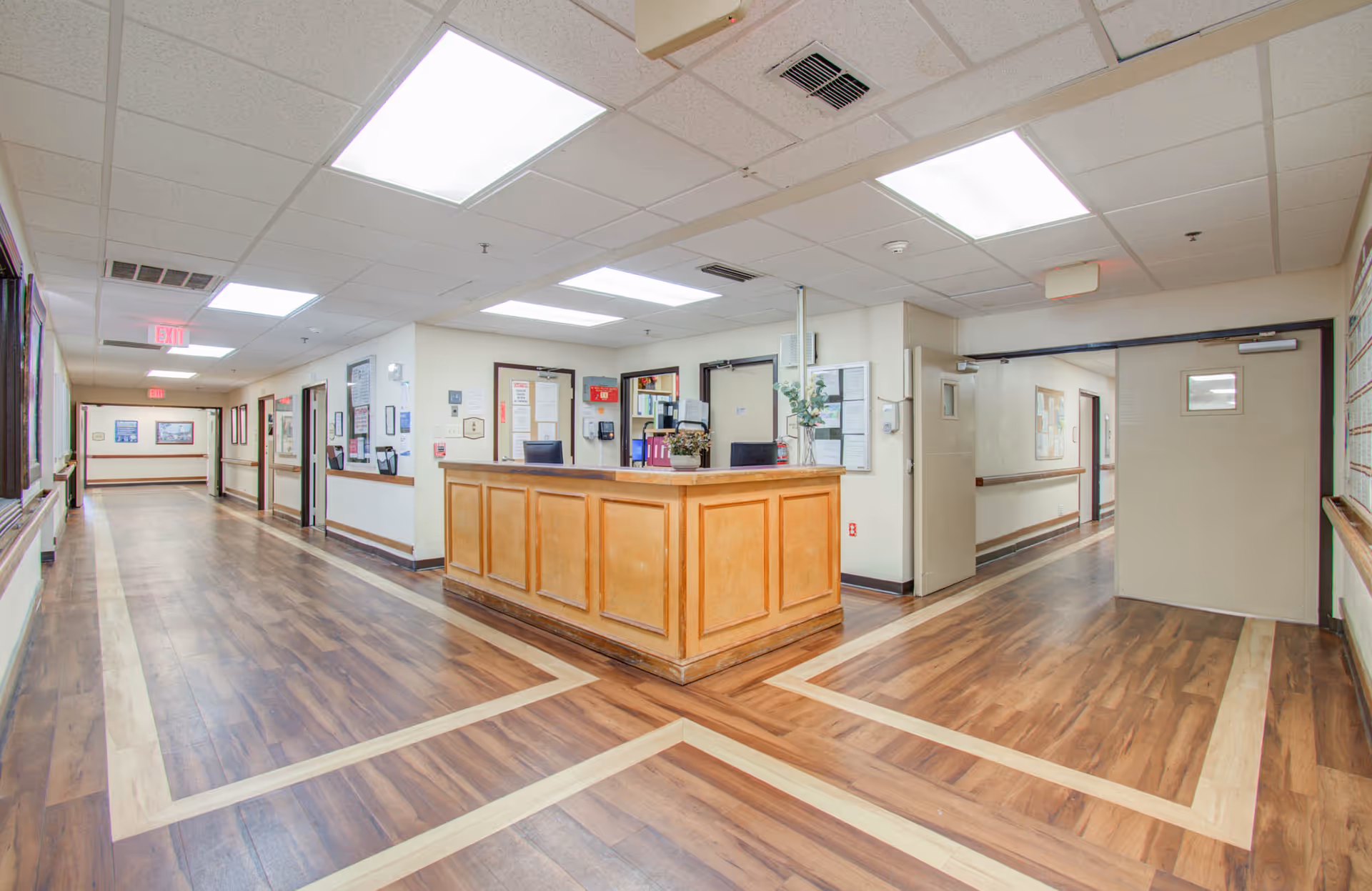 A reception desk in the center of a bright nursing facility hallway with wood-patterned floors, drop ceiling lights, and doors along the corridors.