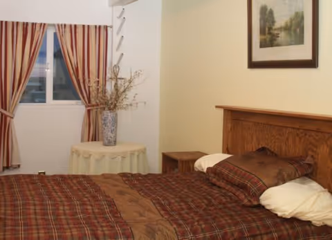 A bedroom with a bed covered in a brown and red plaid comforter and matching pillows. There is a wooden headboard, a small round table with a vase of dried flowers, striped curtains on the window, and a framed landscape painting on the wall.