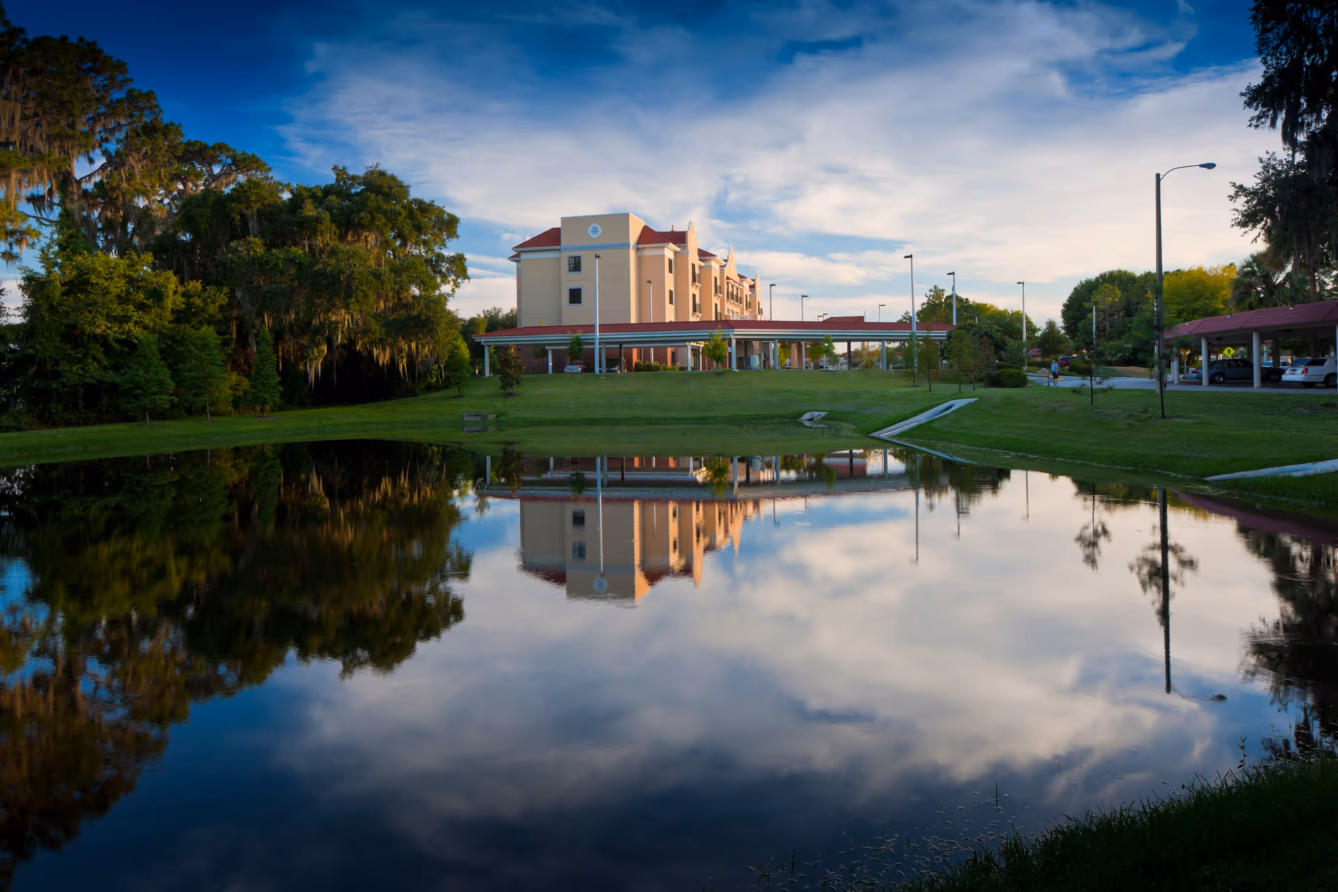 A multi-story residential building reflected in a calm pond with surrounding lawn, trees, and a partly cloudy sky.