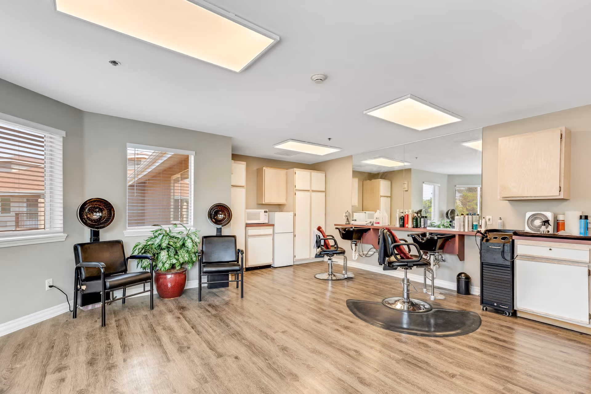 Interior view of a hair salon area in a senior living facility with two black salon chairs in front of a large mirror, hair dryers mounted on stands next to two black waiting chairs, a potted plant between the waiting chairs, and cabinets and a small refrigerator in the background.
