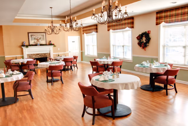 Bright dining room with round tables set for meals, red chairs, chandeliers, windows, and a fireplace.