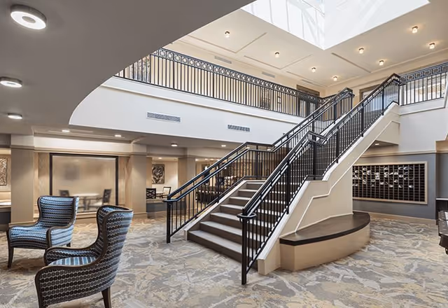 Spacious and well-lit interior of a senior living facility featuring a central staircase with black railings, patterned carpet flooring, and comfortable seating with two upholstered chairs. The area has a high ceiling with a skylight allowing natural light to fill the space, and a second-floor balcony with matching railings overlooking the main floor.