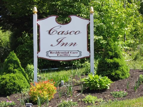 A white wooden sign with gold finials on top of two posts, displaying the text 'Casco Inn Residential Care Facility', surrounded by green shrubs, plants, and a mulched garden bed with grass and trees in the background.