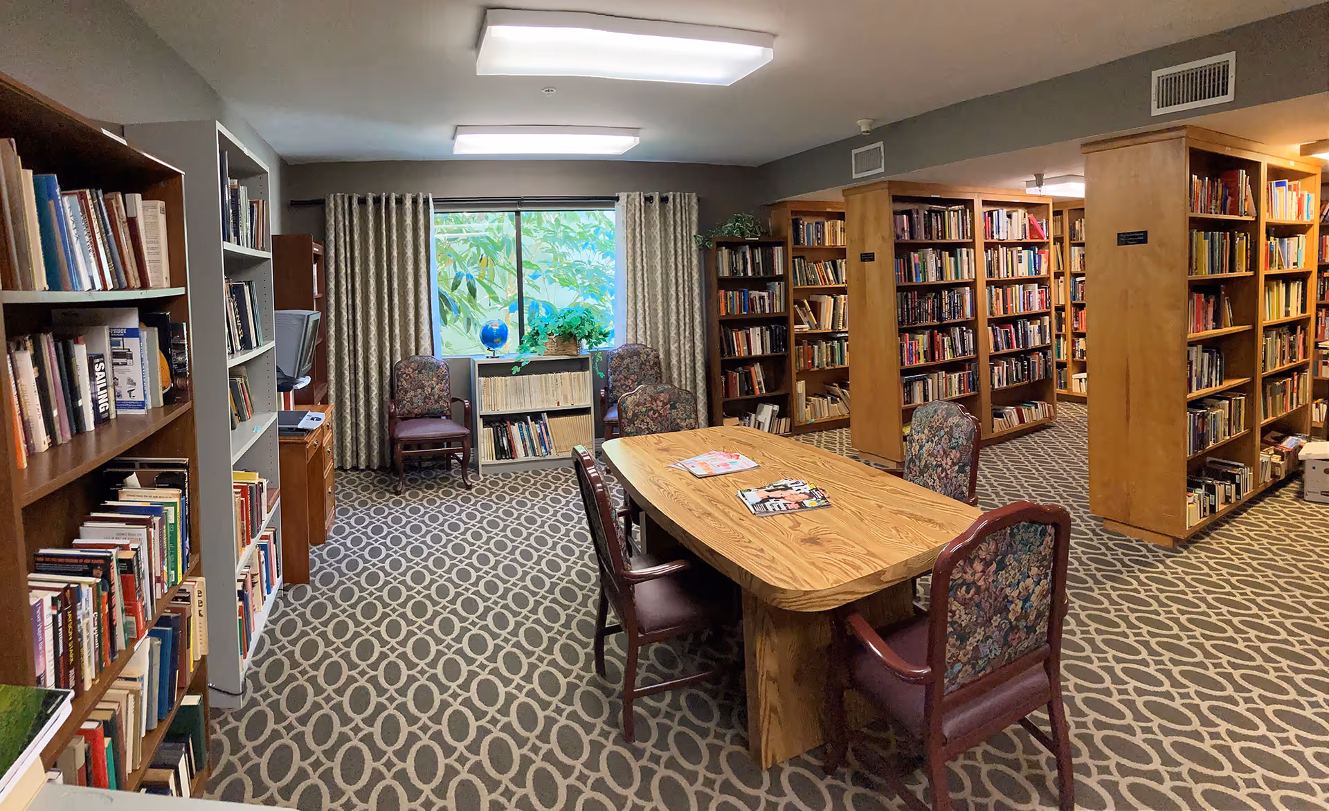 Interior library with bookshelves, a central wooden table surrounded by chairs, patterned carpet, and a window with curtains.