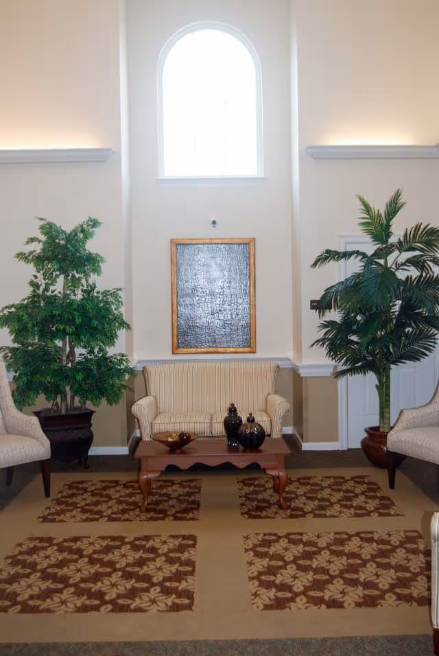 Bright assisted living lobby with a striped loveseat, chairs, coffee table, decorative plants, and a tall arched window.