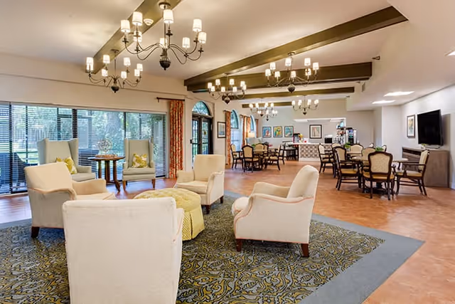 A spacious senior living common area with comfortable armchairs arranged around a patterned rug in the foreground. In the background, there are multiple dining tables and chairs, large windows with blinds, and decorative chandeliers hanging from the ceiling. The room has warm lighting and a welcoming atmosphere.