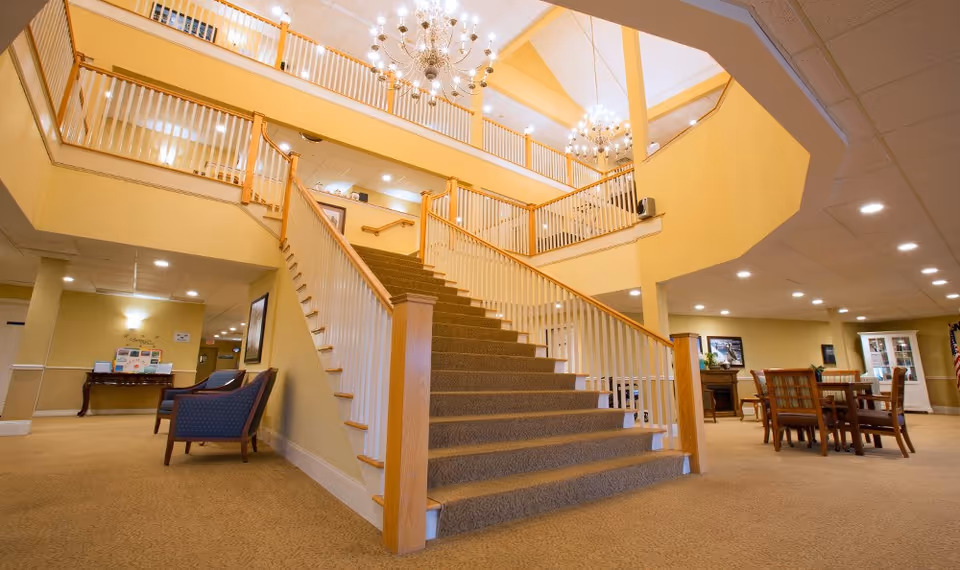 Interior view of a senior living facility featuring a wide carpeted staircase with wooden handrails leading to an upper floor. The area is well-lit with chandeliers and recessed ceiling lights. There are seating areas with chairs and tables on the ground floor, and the walls are painted in warm yellow tones.