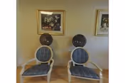 Two vintage-style salon hair drying chairs with blue patterned cushions placed side by side against a beige wall. Above the chairs are two framed floral paintings hanging on the wall.