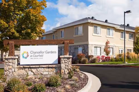Two-story beige senior living building with a stone entrance sign and landscaped surroundings.