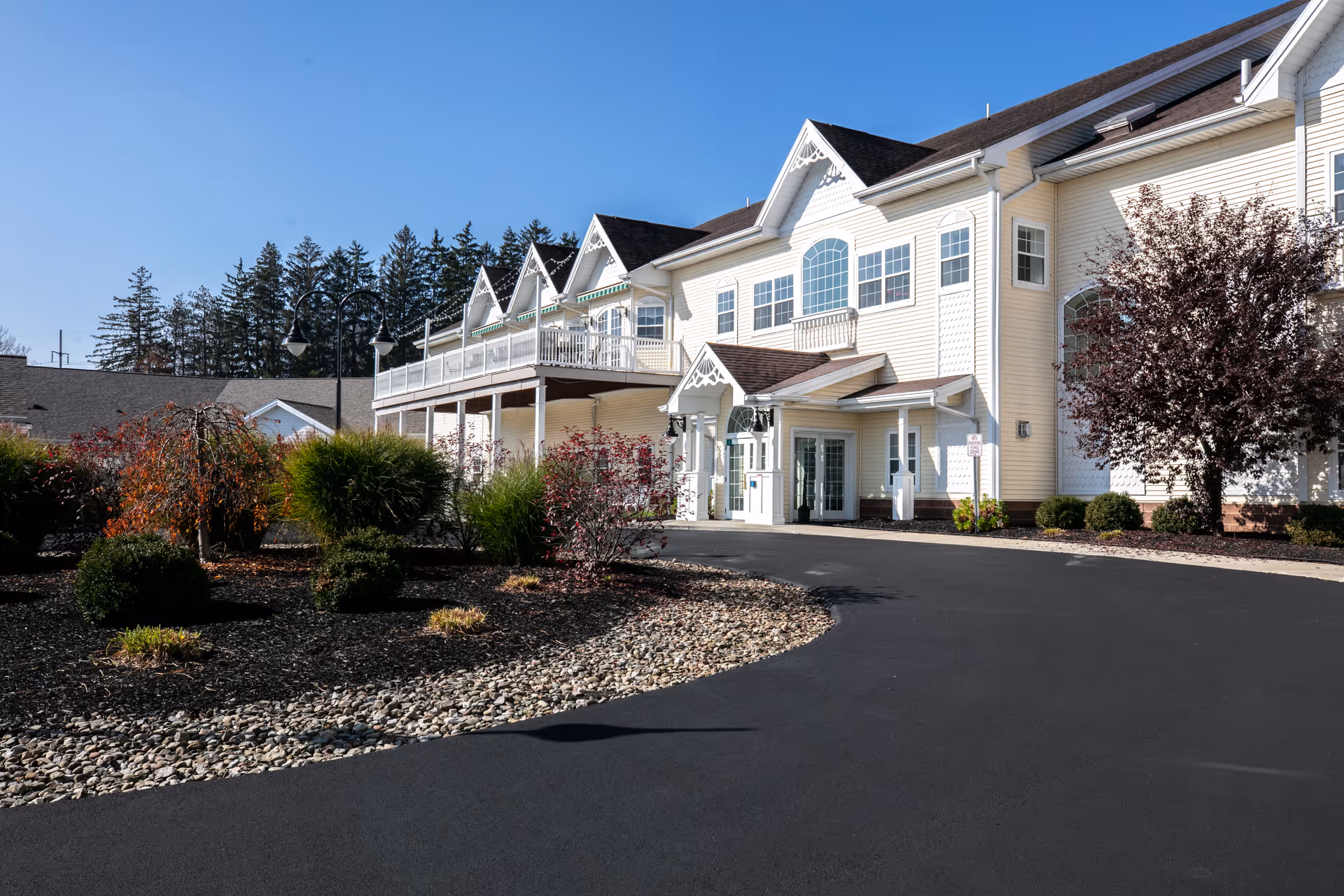 Exterior view of a senior living facility building with white siding, multiple windows, and a covered entrance. The driveway curves in front of the building, and there are landscaped bushes and trees around the area under a clear blue sky.