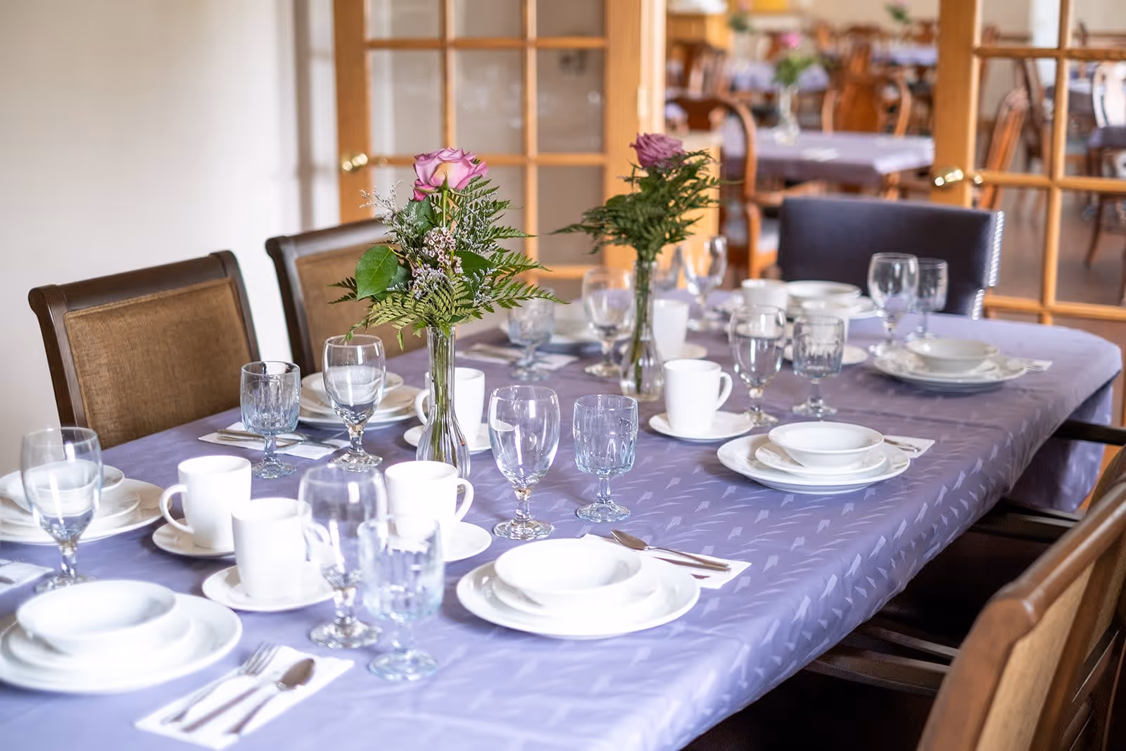A dining table set with white plates, bowls, cups, and various glasses on a lavender tablecloth. There are two small vases with pink roses and greenery as centerpieces. Wooden chairs surround the table, and a glass-paneled door is visible in the background.