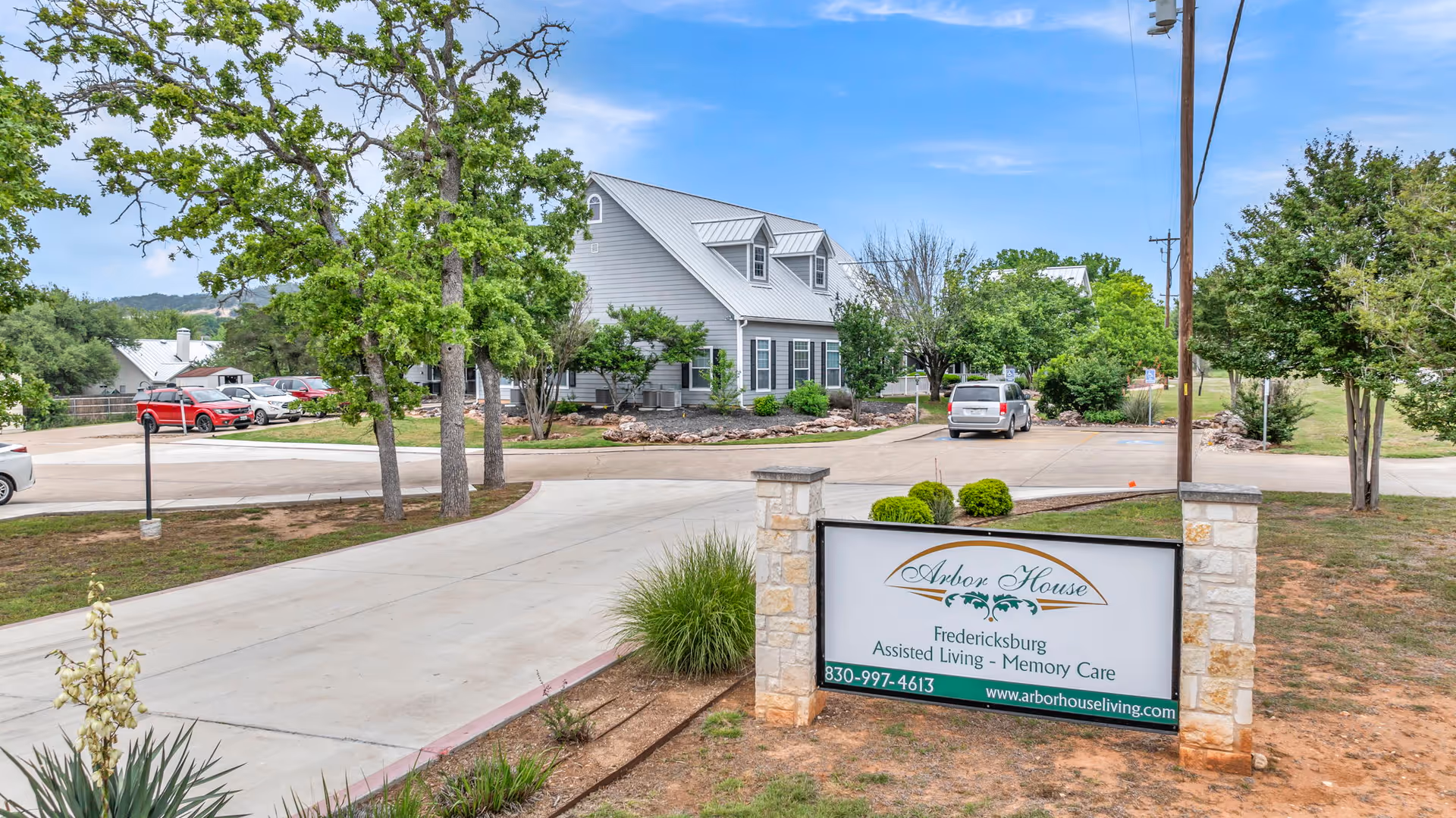 Driveway and entrance sign for Arbor House Fredericksburg assisted living in front of a light-gray two-story building with trees and parked cars.