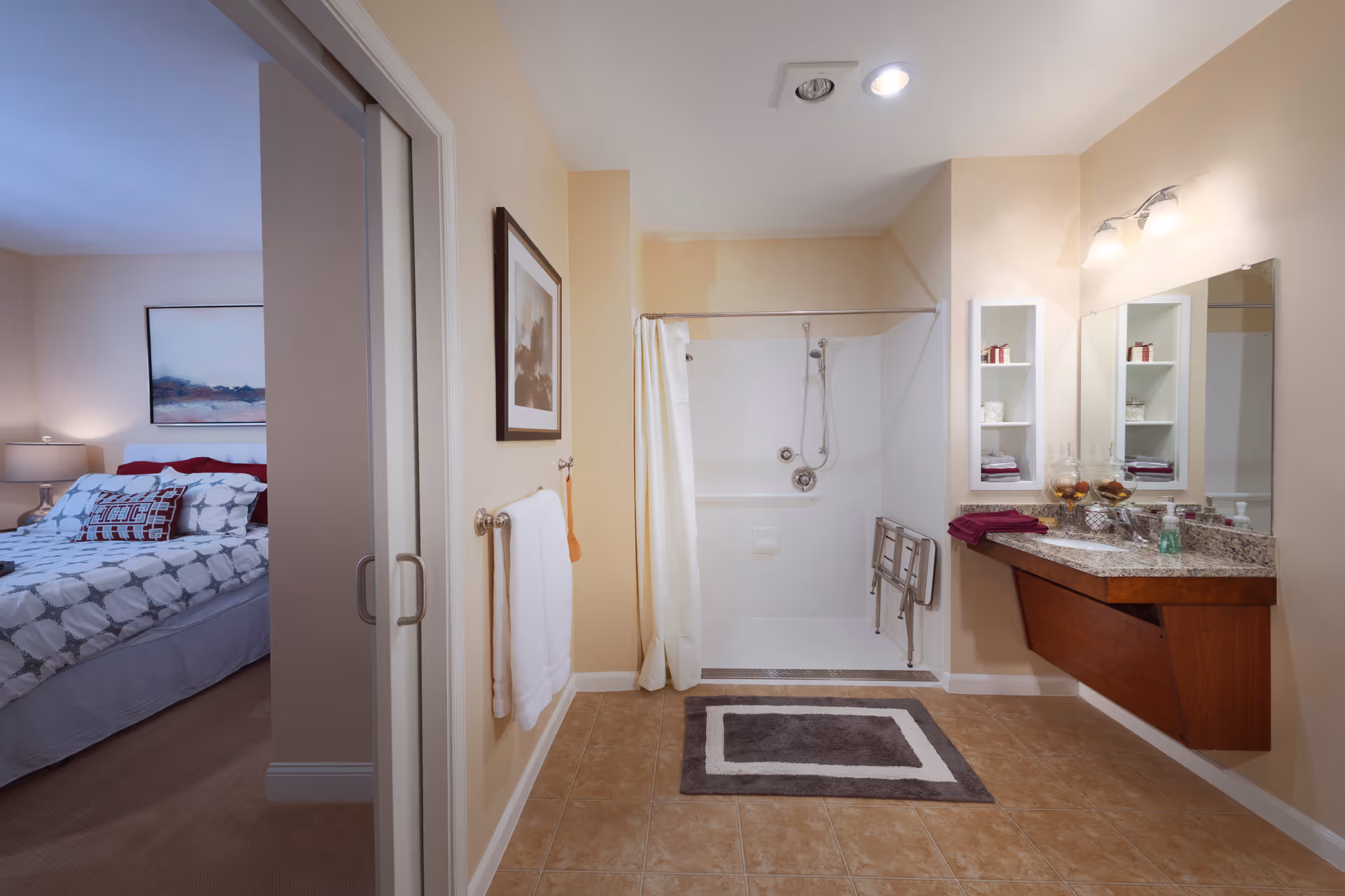 A bathroom with a walk-in shower featuring a grab bar and a shower seat. The bathroom has a large mirror above a granite countertop with a wooden vanity. There are shelves with towels and decorative items. A towel hangs on the wall next to the shower. To the left, a bedroom with a bed, pillows, a bedside lamp, and a framed picture on the wall is partially visible through a sliding door.