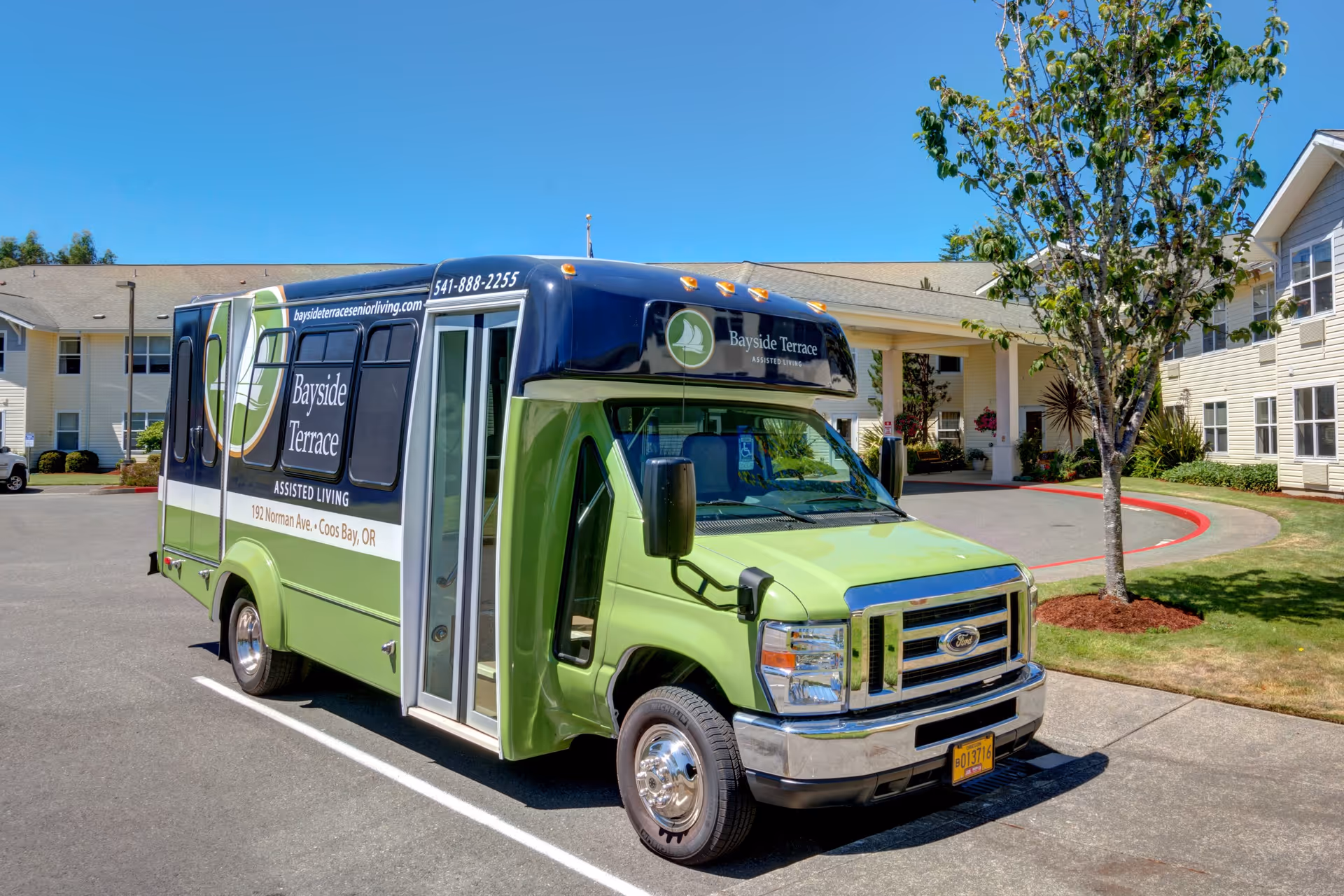 A green and black Bayside Terrace assisted living shuttle bus parked in front of a residential building with a tree and well-maintained landscaping under a clear blue sky.