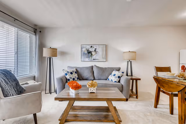 A bright living room with a gray sofa adorned with patterned cushions, a wooden coffee table with two small flower arrangements, a beige armchair with a dark throw, two tall floor lamps, a framed floral painting on the wall, and a wooden dining table with chairs set for a meal.