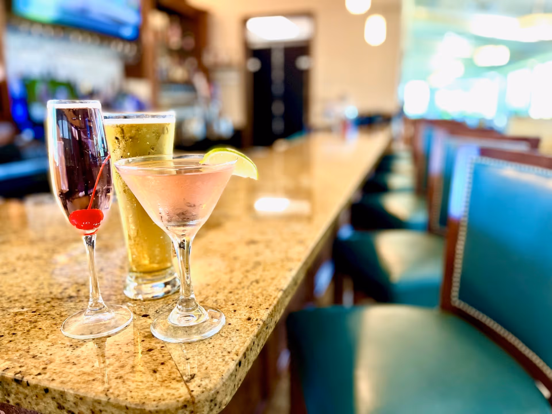 Three drinks on a polished granite bar counter inside a lounge or bar area. The drinks include a champagne flute with a cherry, a tall glass of beer, and a martini glass with a lime wedge. In the background, there are teal cushioned bar stools lined up along the counter and a blurred view of the interior space with warm lighting.