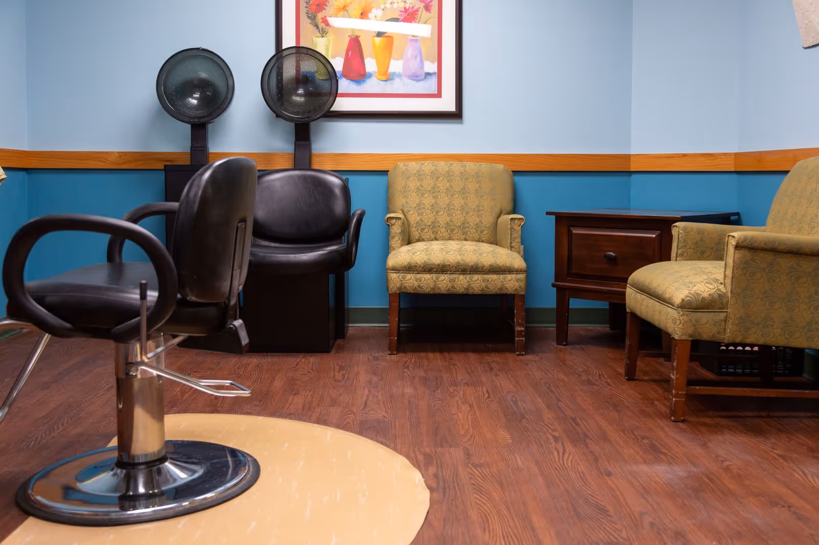 Interior room with two black salon hair dryer chairs, a black salon styling chair, two patterned armchairs, a wooden side table, and a colorful framed painting on the wall. The walls are painted blue with a wooden trim and the floor has a wood finish.