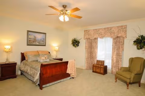 A cozy bedroom in an assisted living facility featuring a wooden bed with beige and green bedding, two bedside tables with lamps, a green armchair, a window with floral curtains, and decorative plants on the walls.