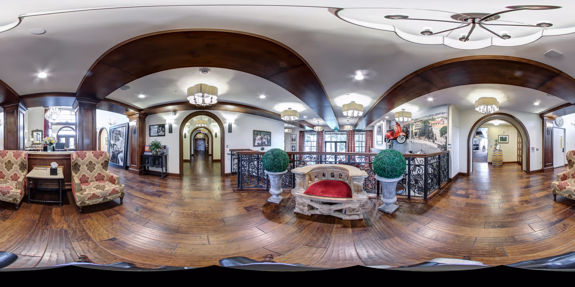 A spacious and elegant interior common area with wooden floors and dark wood ceiling beams. The room features patterned upholstered chairs, a decorative wooden bench with a red cushion, two large potted plants, and ornate light fixtures. There are arched doorways and a hallway leading further into the building. The walls are adorned with framed pictures and a large mural with a red scooter mounted on it.