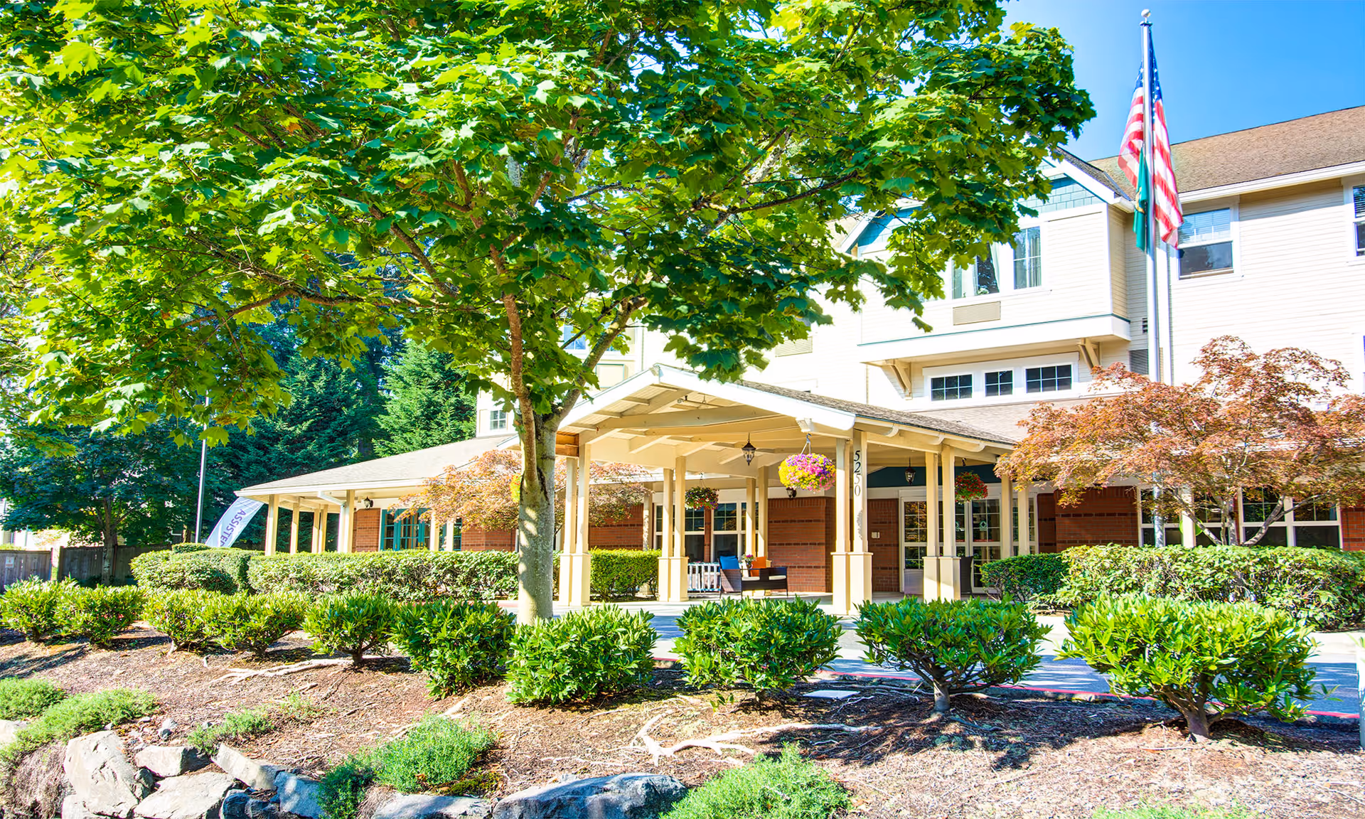 Front exterior view of The Village of St. Edward at Green senior living facility with a covered entrance, surrounded by green trees and shrubs under a bright sunny sky.