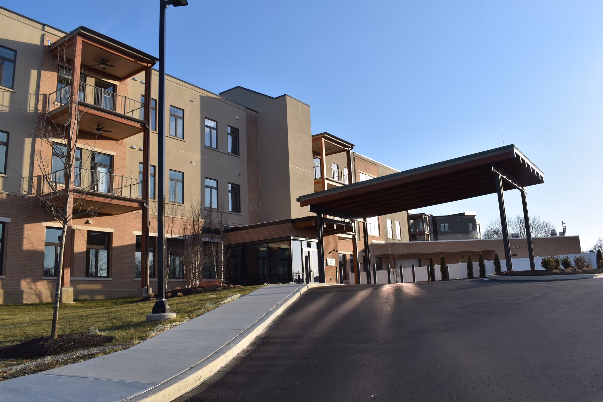 Exterior view of a multi-story senior living facility building with balconies, large windows, and a covered entrance driveway under a clear blue sky.