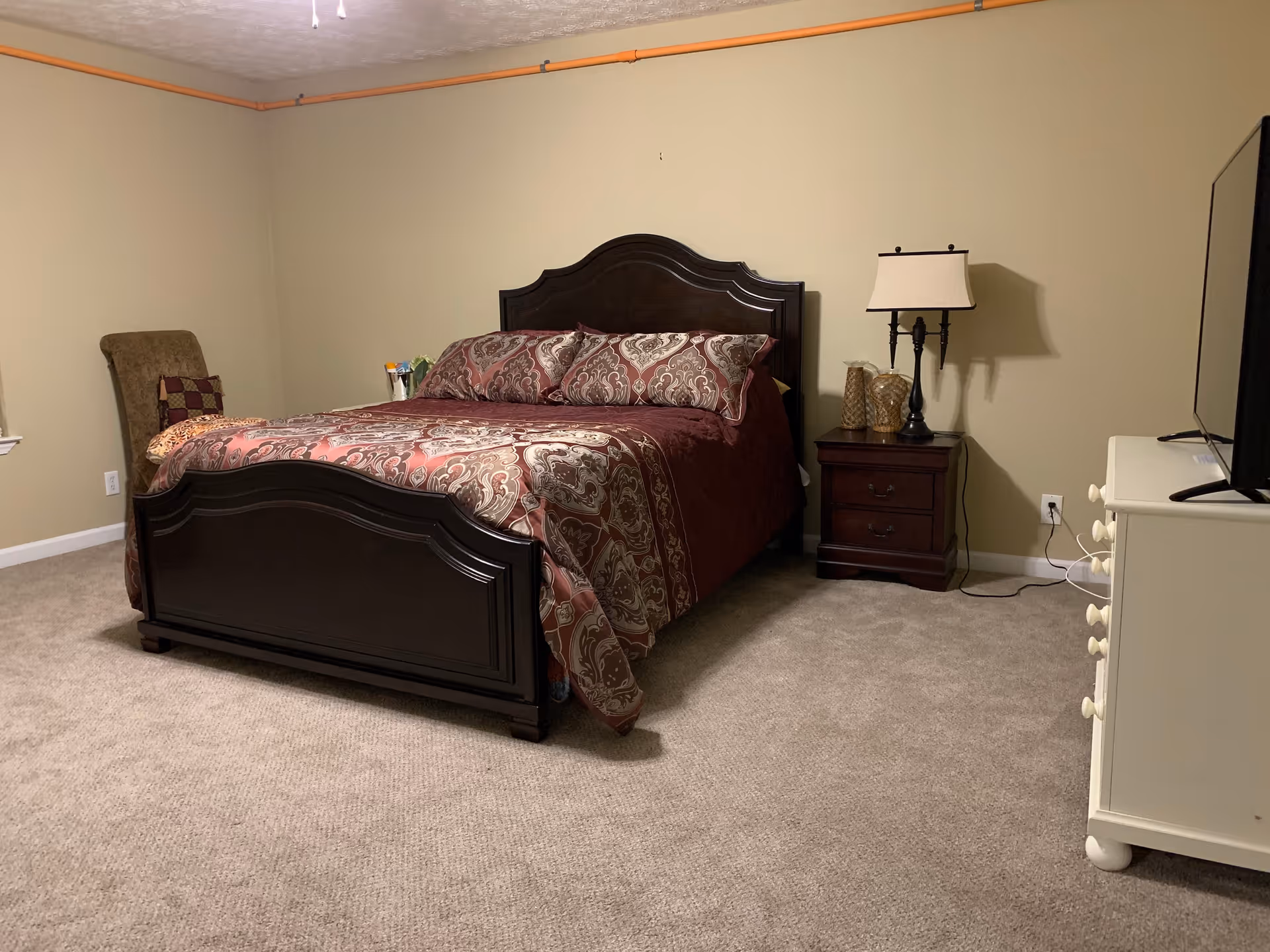 A bedroom with a dark wooden bed frame and matching headboard, covered with a patterned maroon and beige bedspread and pillows. To the right of the bed is a dark wooden nightstand with a lamp and decorative items. A white dresser with a TV on top is positioned on the right side of the room. The walls are beige, and the floor is carpeted in a light brown color. There is also a cushioned chair with a pillow in the left corner of the room.