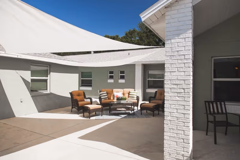 Outdoor patio area with cushioned wicker chairs and a sofa arranged around a coffee table, shaded by white fabric canopies attached to the building. The building walls are light gray with several windows, and the sky is clear and blue.