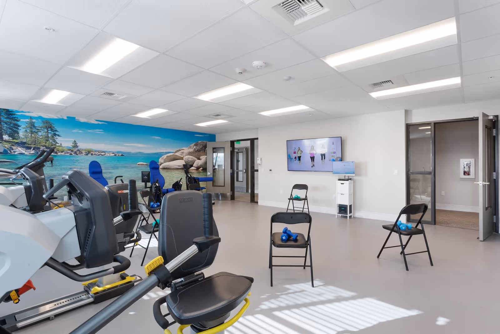 A spacious exercise room with various fitness equipment including stationary bikes and chairs with dumbbells placed on them. A large mural of a serene lake with rocks and trees covers one wall. A flat-screen TV mounted on the opposite wall displays an exercise video with three people working out. The room has bright overhead lighting and a clean, modern design.