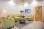 A cozy seating area in a health and rehabilitation center featuring a green sofa, two matching green armchairs, a round wooden coffee table with a flower arrangement, a floor lamp, a potted plant, and a wall-mounted television. The room has tiled flooring, a patterned rug, and a wooden door with a window.