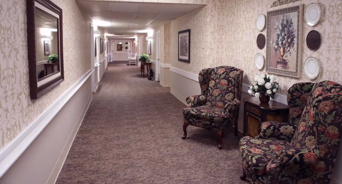 A long, carpeted hallway in an assisted living facility with floral-patterned armchairs and a small wooden table with a vase of white flowers on the right side. The walls have a beige patterned wallpaper with framed artwork and decorative plates. Mirrors and small tables with plants line the left side of the hallway.