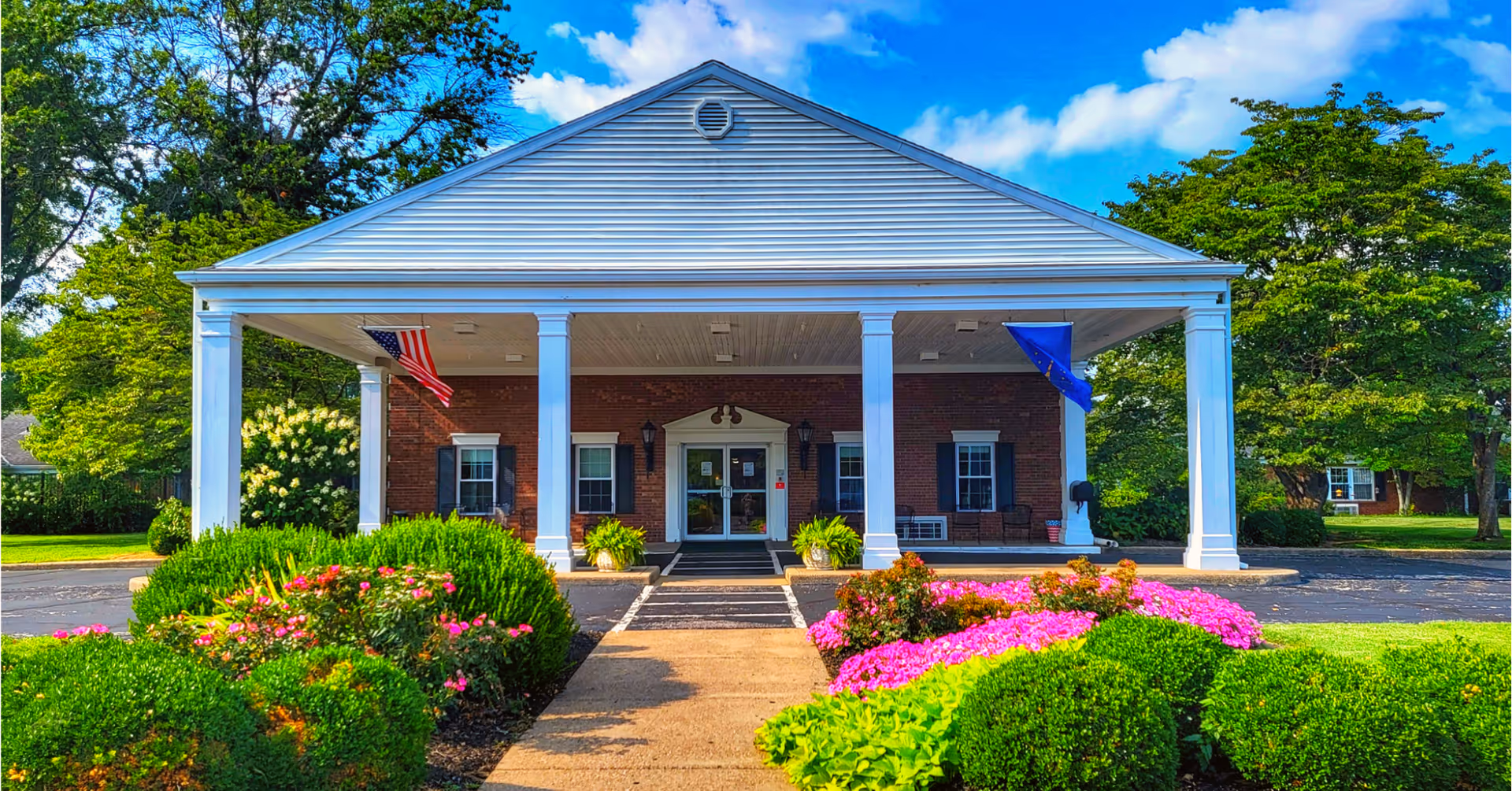 Front exterior view of a brick building with white columns and a covered entrance. There are two flags hanging under the roof, one American flag and one blue flag. The walkway leading to the entrance is bordered by well-maintained bushes and colorful flowers. Trees and greenery surround the building under a partly cloudy blue sky.