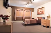Spacious furnished bedroom with a bed, nightstands, dresser, mounted TV, and a window with blinds and curtains.