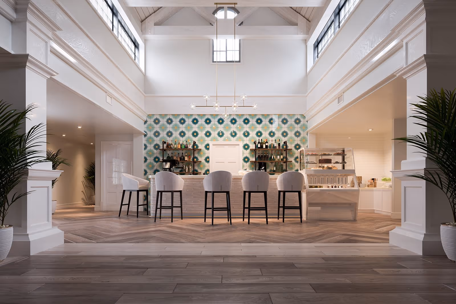 A bright and spacious interior area featuring a modern bar with five cushioned bar stools. The back wall has a colorful geometric patterned wallpaper with shelves holding bottles and glassware. To the right, there is a display case with pastries and other food items. The room has high ceilings with large windows near the top, letting in natural light, and two large potted plants on either side of the entrance.