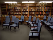 Small library or meeting room with rows of upholstered chairs facing forward and bookshelves covering the back wall.