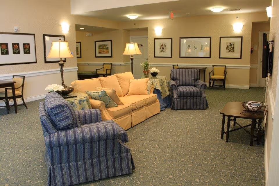 A furnished communal sitting room with a yellow sofa, blue striped armchairs, side tables, lamps, framed artwork, and a wall-mounted TV.