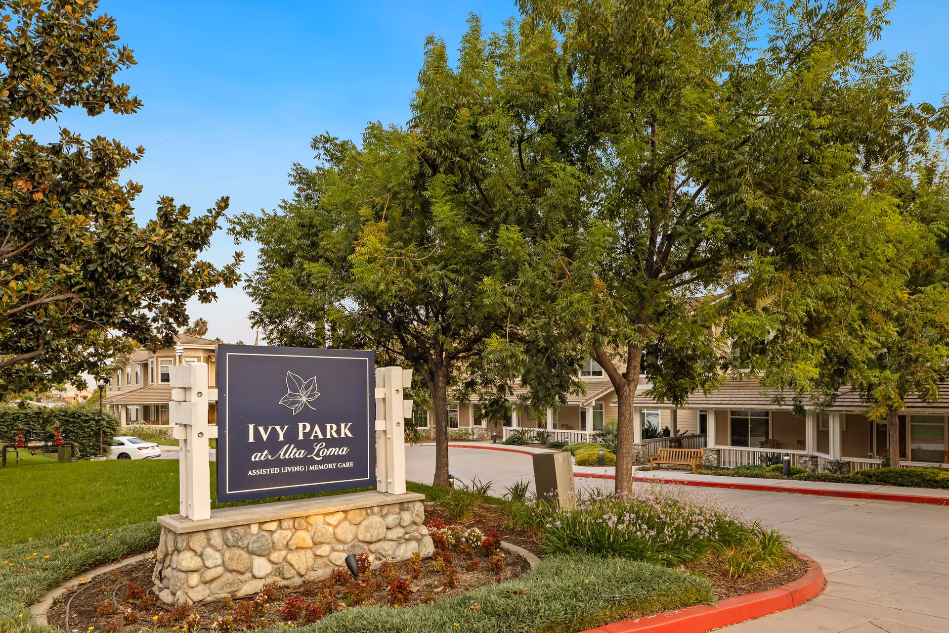 Entrance sign for Ivy Park at Alta Loma assisted living and memory care facility, surrounded by trees and landscaping with a building visible in the background under a clear blue sky.