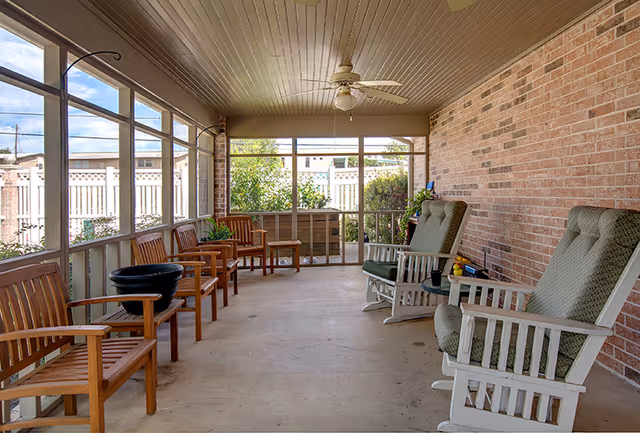 A screened-in porch area with wooden chairs lined up along the left side and two cushioned rocking chairs on the right side against a brick wall. The porch has a ceiling fan and overlooks a fenced yard with greenery outside.