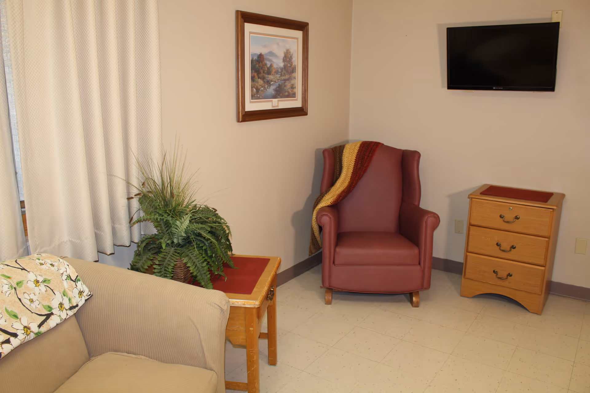 A cozy corner of a room featuring a beige couch with a floral blanket, a wooden side table with a potted green plant, a maroon armchair with a multicolored knitted throw draped over it, a wooden three-drawer nightstand with a red mat on top, a wall-mounted flat-screen TV, and a framed landscape painting on the wall.