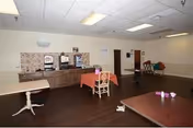 A spacious room with dark wood flooring and white walls featuring a beverage station with coffee machines and cups along one wall. There are a few tables with chairs, some covered with orange tablecloths, and a few scattered items on the floor. The ceiling has recessed lighting and a wall-mounted light fixture.