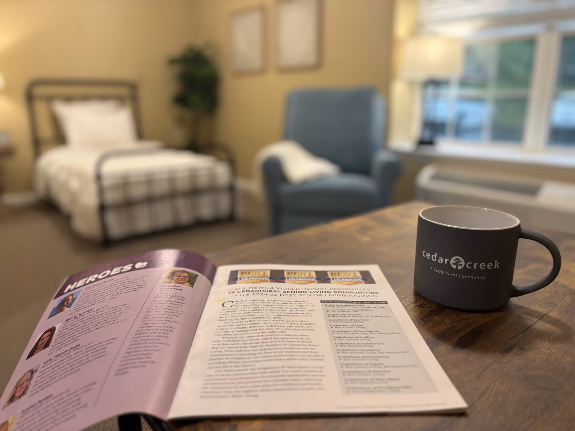 A cozy assisted living room with a single bed in the background, a blue armchair, and a wooden table in the foreground. On the table, there is an open magazine and a black coffee mug with the text 'cedar creek A Cedarhurst Community'. The room has warm lighting and a window with a view outside.