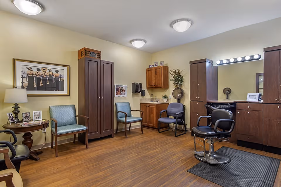 Interior of a salon room in a senior living facility with wooden flooring, beige walls, and ceiling lights. The room contains salon chairs, a large mirror with lights above it, wooden cabinets, a small sink area, two blue cushioned chairs, a side table with a lamp and framed photos, and wall decorations including a framed picture of women in black dresses and hats.